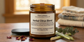 An amber glass jar of Pure Lumens Herbal Citrus Blend essential oil and soy wax candle sits on a wooden table with a lid, lemongrass stems, and dried petals in the foreground, and folded towels blurred in the background.